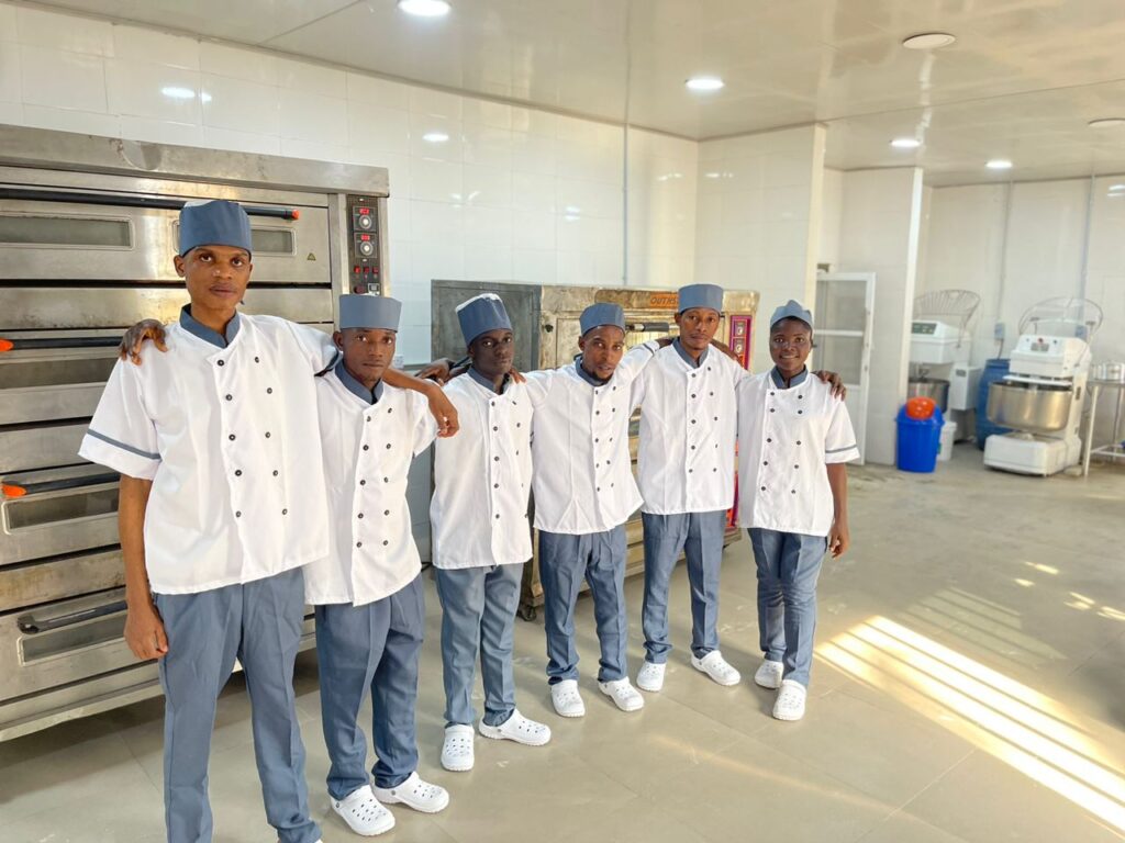 Six apprentice bakers in matching uniforms pose beside commercial ovens inside bakery.
